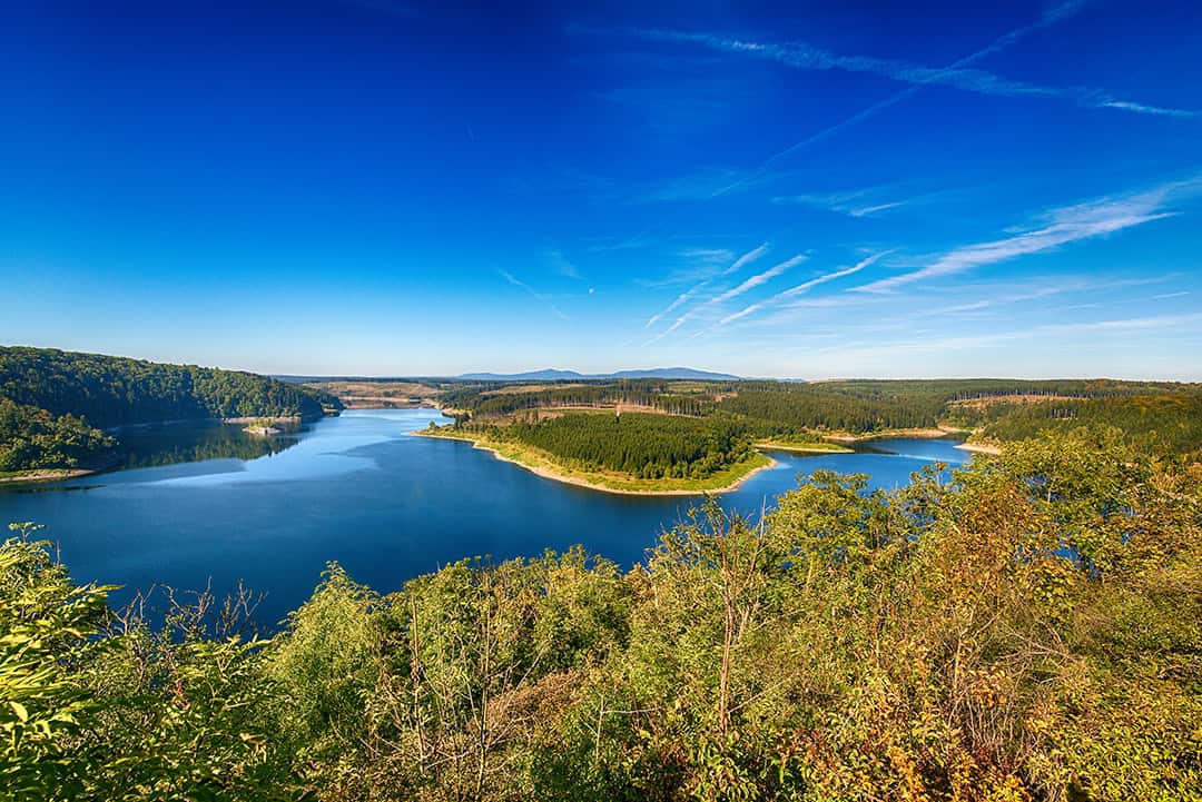 Ein malerischer Blick auf einen von grünen Wäldern und Hügeln umgebenen See unter einem klaren blauen Himmel mit Wolkenfetzen, gesehen von einem erhöhten Aussichtspunkt mit Bäumen im Vordergrund.