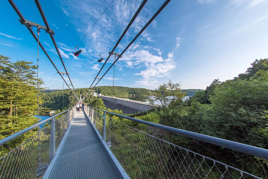 Eine lange Hängebrücke aus Metall führt über einen üppigen, grünen Wald, in dessen Hintergrund ein Damm und ein See zu sehen sind. Menschen gehen auf der Brücke spazieren, und eine Person fährt auf einer Seilrutsche über dem blauen Himmel mit vereinzelten Wolken.