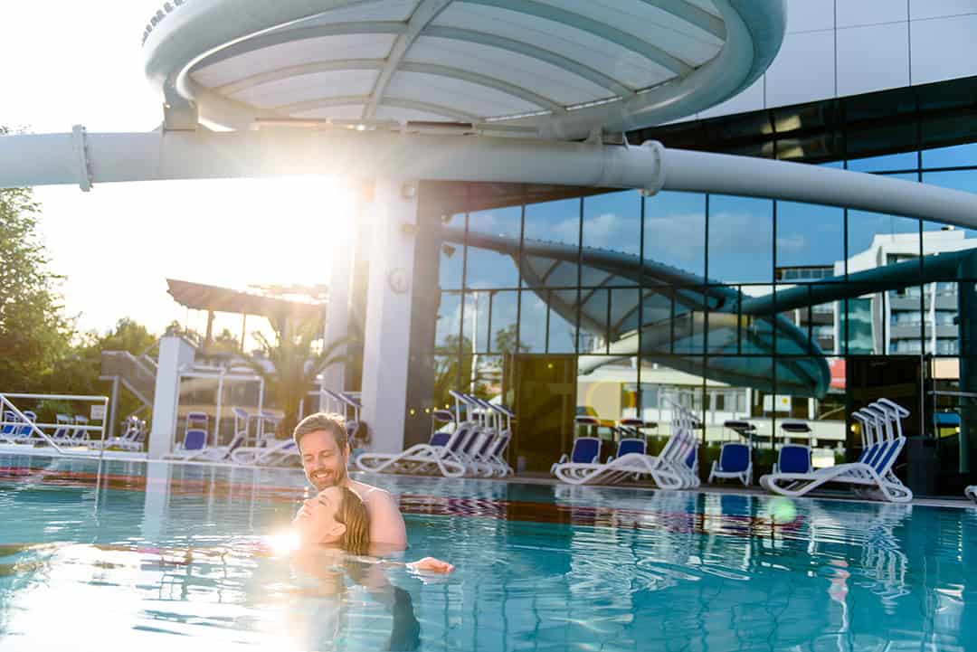 BFÜ-2019-Europa-Therme_1080 Ein Mann und ein Kind lächeln, während sie an einem sonnigen Tag zusammen in einem Freibad schwimmen, mit modernen Gebäuden und leeren Liegestühlen im Hintergrund. Das Sonnenlicht wird vom Wasser reflektiert und schafft eine helle, fröhliche Szene.