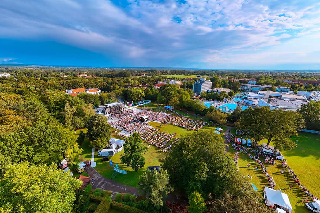 BR-Schlagerparty_OpenAir-2024©Christoph_Braml_1080 Luftaufnahme eines Musikfestivals im Freien mit einer großen Menschenmenge, einer Bühne und Zelten in einem grünen Park, umgeben von Bäumen, Gebäuden und Teichen unter einem teilweise bewölkten Himmel.