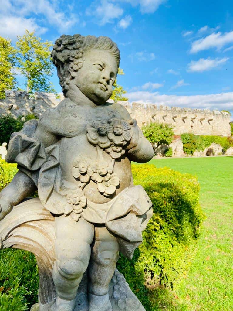 Eine mit Blumen geschmückte Steinstatue eines engelsgleichen Kindes steht in einem sonnenbeschienenen Garten mit grünem Gras, ordentlich gestutzten Büschen und einer alten Steinmauer im Hintergrund unter einem blauen Himmel mit vereinzelten Wolken.