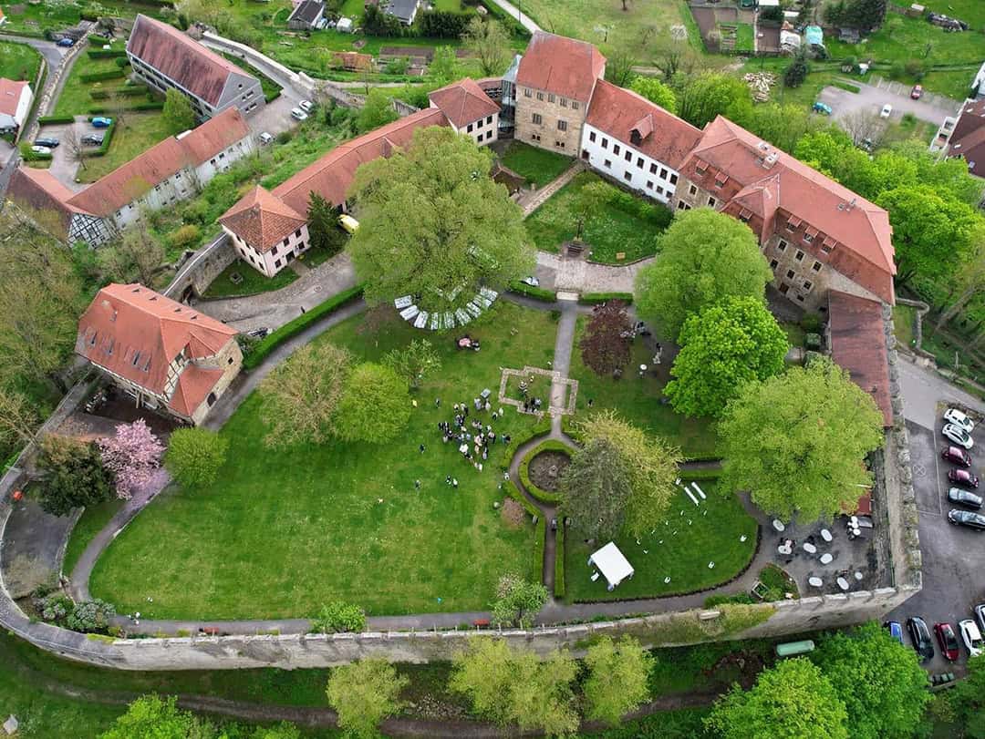 Luftaufnahme eines historischen Gebäudekomplexes mit roten Dächern, die einen großen, grünen Innenhof umgeben, in dem sich Menschen versammeln. Auf dem Gelände und in der Umgebung sind Bäume, Gärten und geparkte Autos zu sehen.