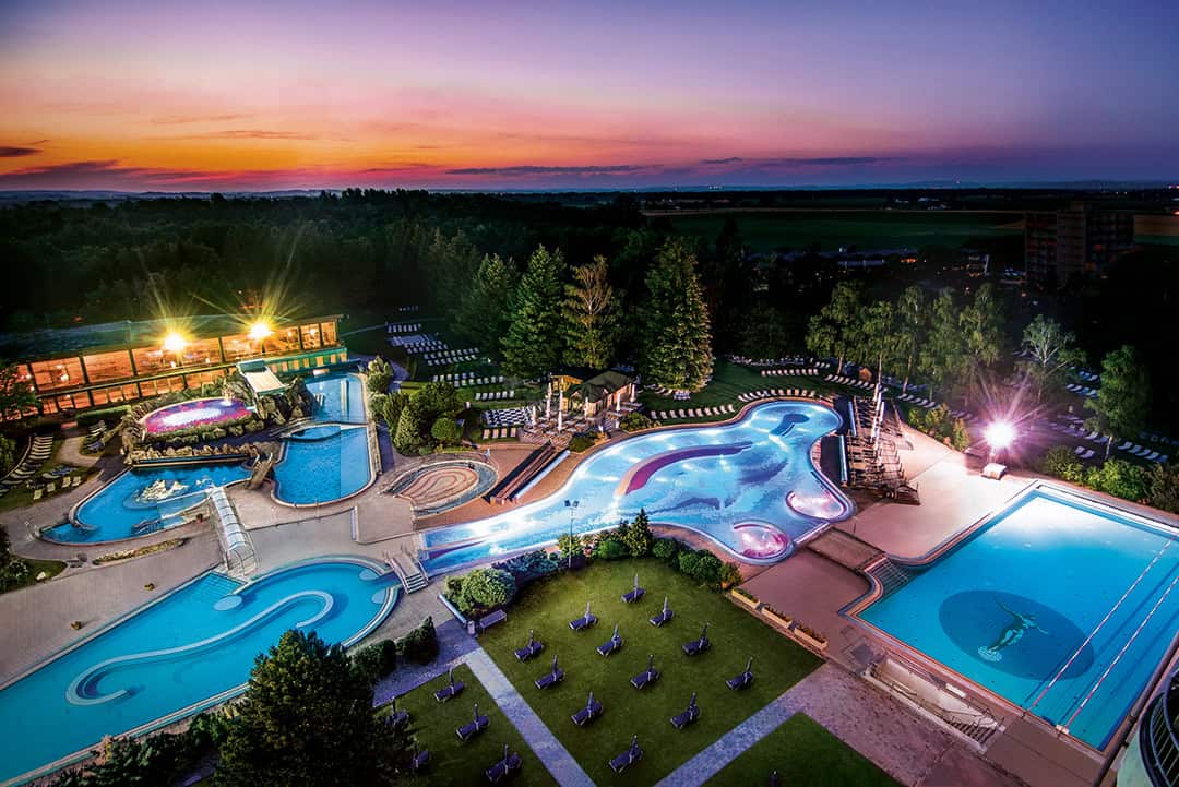 Johannesbad-Therme_1080 Ein großer, beleuchteter Außenpoolkomplex bei Sonnenuntergang mit miteinander verbundenen Becken, einem Sprungbrett, Liegestühlen und umliegenden Bäumen, mit einem leuchtenden Himmel im Hintergrund.