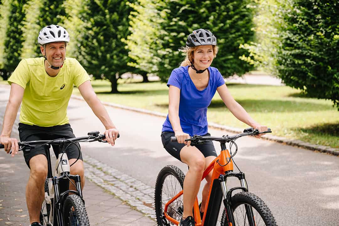 Radfahren©LeonieLorenz-4659_1080 Zwei Personen mit Helmen fahren an einem sonnigen Tag mit dem Fahrrad auf einem gepflasterten Weg durch einen grünen Park. Beide lächeln; die Person auf der linken Seite trägt ein gelbes Hemd, die Person auf der rechten Seite ein blaues Hemd und fährt ein orangefarbenes Fahrrad.
