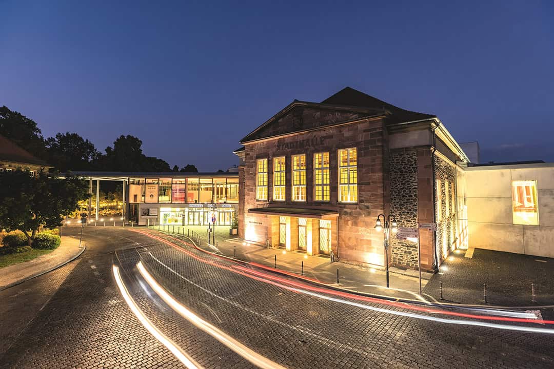 Ein historisches Backsteingebäude mit großen Fenstern wird in der Abenddämmerung beleuchtet, wobei sich die Lichtspuren der vorbeifahrenden Fahrzeuge durch die Kopfsteinpflasterstraße davor schlängeln. Im Hintergrund sind Bäume und ein moderner Anbau zu sehen.