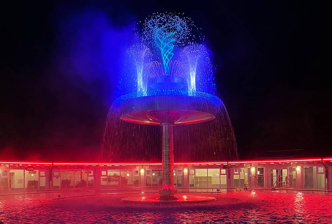 Therme_Eins_Schwammerl_1080 Ein mehrstöckiger Springbrunnen wird nachts mit leuchtenden blauen und roten Lichtern beleuchtet. Das Wasser spritzt von oben und erzeugt einen bunten Nebel, während das umliegende Gebäude in rotes Licht getaucht wird.