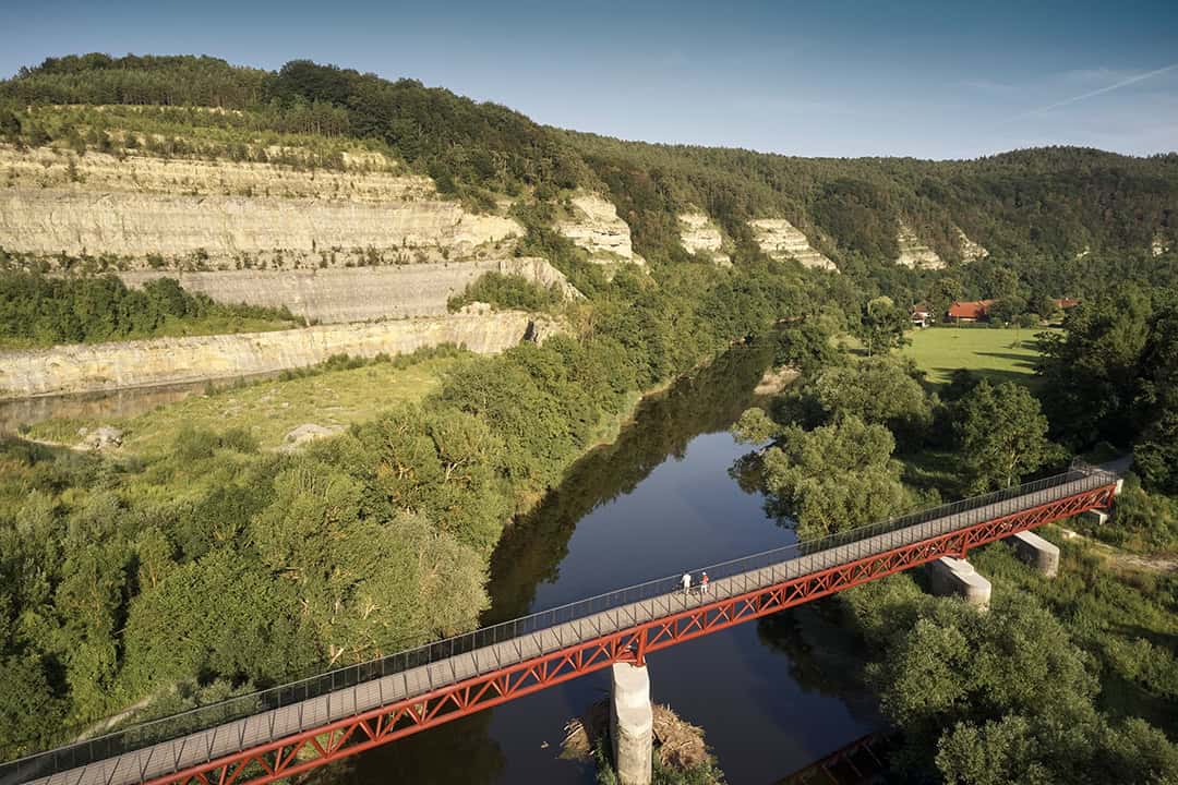 Eine rote Fußgängerbrücke überquert einen Fluss, umgeben von üppigem Grün. Im Hintergrund sind terrassierte Klippen und bewaldete Hügel zu sehen, unter einem klaren blauen Himmel. Zwei Menschen gehen über die Brücke.