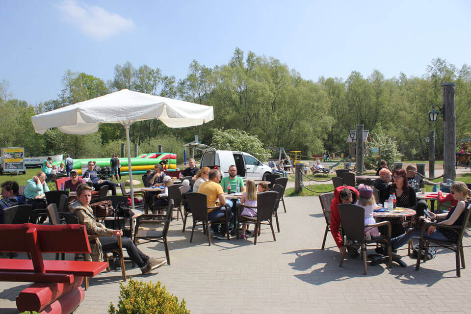 An einem sonnigen Tag sitzen Menschen an Tischen im Freien, essen und unterhalten sich. Ein weißer Regenschirm spendet Schatten. Im Hintergrund sind grünes Gras, Bäume, Spielgeräte und spielende Kinder zu sehen.