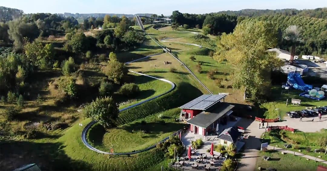 Eine gewundene alpine Rutschbahn schlängelt sich durch eine grüne, grasbewachsene Landschaft mit Bäumen und Hügeln. Ein kleines Gebäude und Sitzgelegenheiten im Freien sind im Vordergrund neben der Bahn zu sehen. Die Szene ist hell und sonnig.