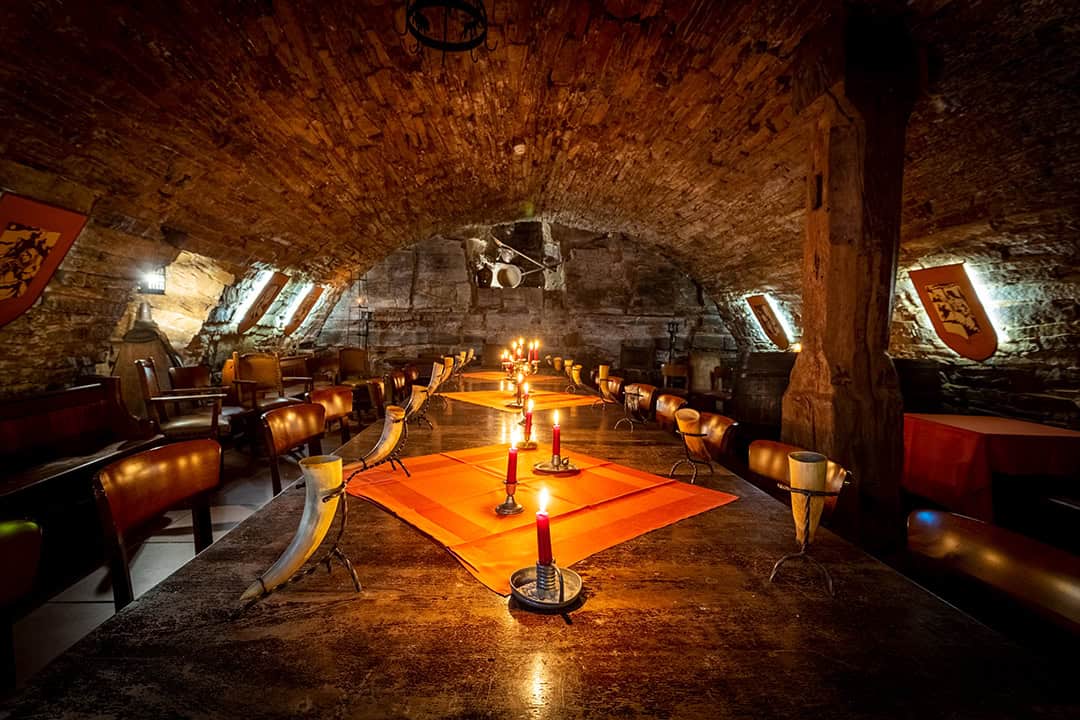 Ein schwach beleuchteter, mittelalterlich anmutender Speisesaal mit Steinwänden und gewölbter Decke, langen, mit Kerzen gedeckten Tischen, orangefarbenen Tischläufern, Trinkhörnern und Wappen an den Wänden.