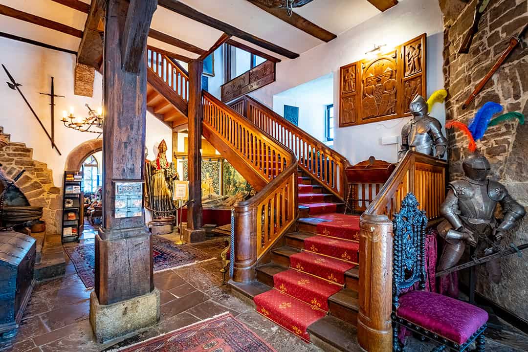 Eine große Treppe in einem mittelalterlichen Interieur mit Holzbalken, Steinwänden, rotem Teppich, Rüstungen, alten Waffen, verzierten Teppichen und dekorativen Tafeln, die eine burgähnliche Atmosphäre schaffen.