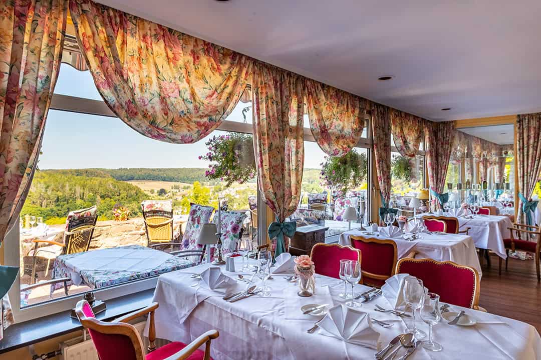 Eleganter Restaurantbereich mit roten Stühlen, weißen Tischtüchern, Gläsern und Servietten. Große Fenster mit geblümten Vorhängen bieten einen Blick auf eine malerische Landschaft, und Topfblumen sorgen für eine gemütliche, einladende Atmosphäre.