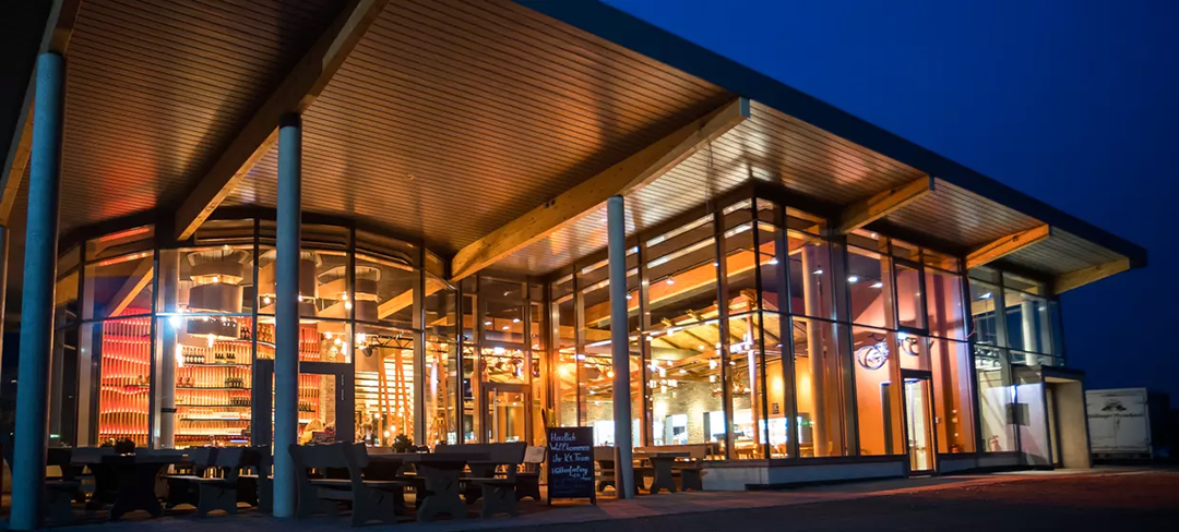 Ein modernes Restaurant mit großen Glasfenstern und Holzbalken wird nachts von innen warm beleuchtet und bietet ein einladendes Interieur mit Tischen, Stühlen und einem leuchtenden Ambiente.