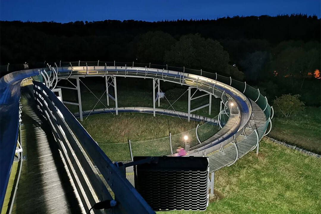 Eine geschwungene, erhöhte Metallrutsche mit Sicherheitsnetzen ist nachts beleuchtet und schlängelt sich durch eine von Bäumen umgebene Rasenfläche unter einem dunklen Himmel.