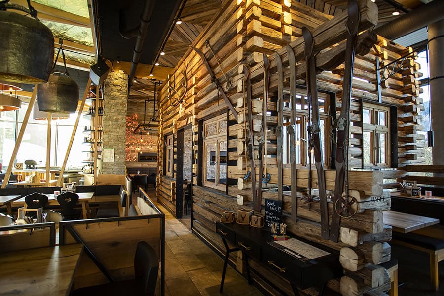 Ein gemütliches Restaurant mit Holzwänden im Blockhausstil, alten Werkzeugen als Dekoration, großen Fenstern und ordentlich angeordneten Holztischen und -stühlen im ganzen Raum. Die warme Beleuchtung schafft eine einladende Atmosphäre.