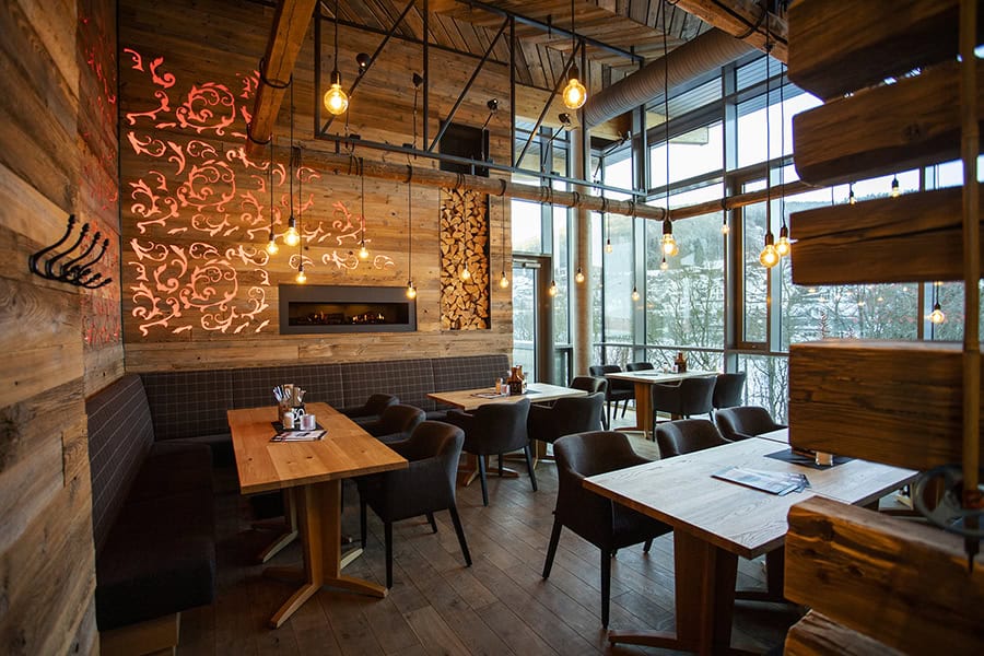 Ein gemütliches Restaurant mit Holzwänden und -böden, modernen schwarzen Stühlen und hängenden Edison-Lampen. Große Fenster lassen natürliches Licht herein, und dekorative Muster zieren eine Wand über einem eingebauten Kamin.