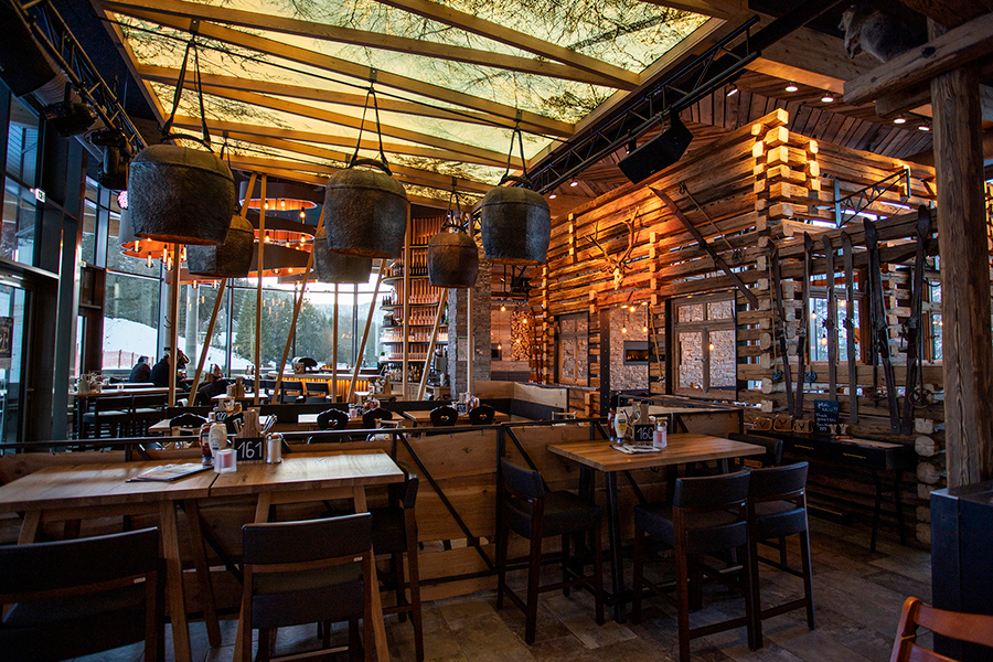 Ein rustikales Restaurant mit Holztischen und -stühlen, warmer Beleuchtung, hängenden Metallkorblampen und Wänden aus Holzlatten schafft eine gemütliche, einladende Atmosphäre. Große Fenster lassen viel natürliches Licht herein.