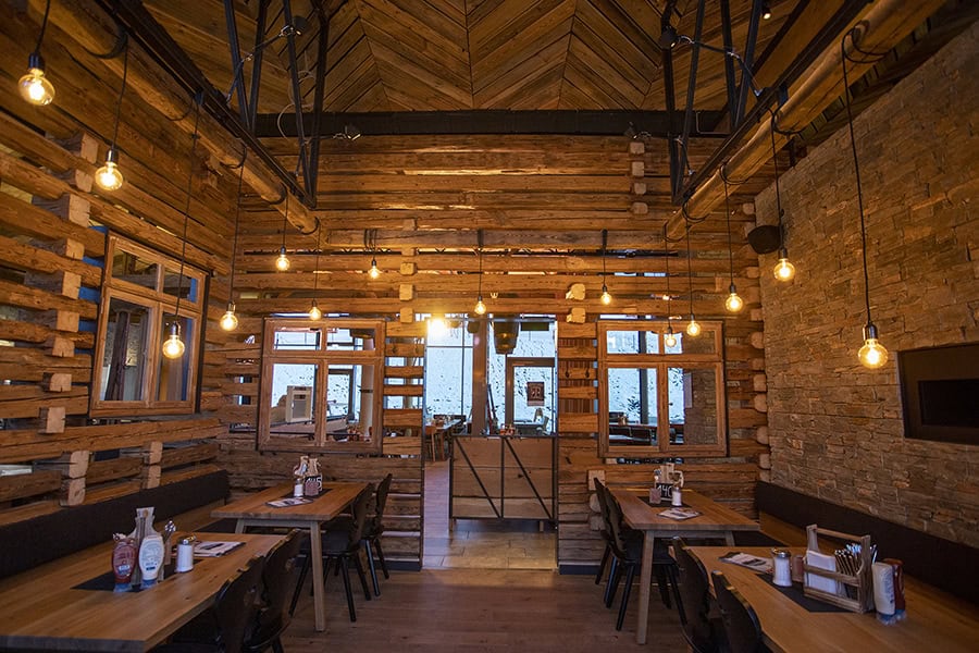 Ein gemütliches Restaurant mit Wänden aus Holzbalken, hängenden Glühbirnen, ordentlich gedeckten Tischen und großen Fenstern, die natürliches Licht hereinlassen. Die Einrichtung hat eine rustikale, warme und einladende Atmosphäre.