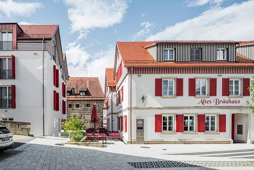 Ein Hof mit modernen weißen Gebäuden mit roten Fensterläden und Ziegeldächern; ein Gebäude trägt ein Schild mit der Aufschrift Altes Bräuhaus, und draußen sind rote Terrassenschirme unter einem teilweise bewölkten Himmel aufgestellt.