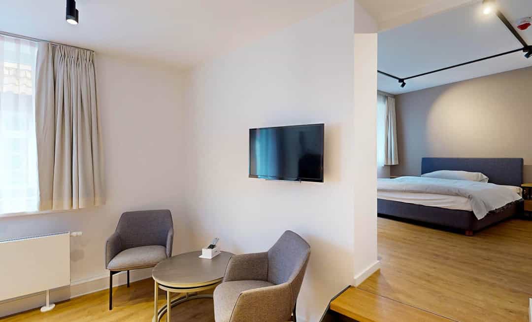 Modernes Hotelzimmer mit Holzboden, einem an der Wand montierten Flachbildfernseher, zwei grauen Sesseln und einem kleinen runden Tisch. Ein Doppelbett ist in einem separaten Schlafbereich durch eine offene Tür sichtbar.