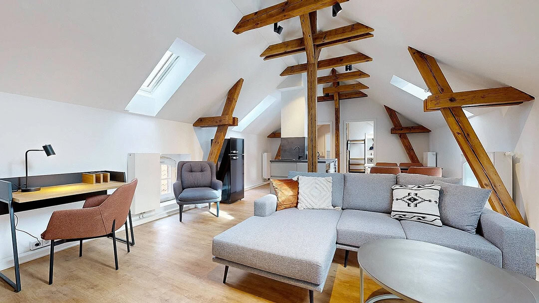 Moderner Wohnraum im Dachgeschoss mit freiliegenden Holzbalken, einem grauen Sektionssofa, einem runden Couchtisch, einem Schreibtisch mit Stuhl und Dachflächenfenstern, die natürliches Licht auf den Holzboden fallen lassen. Die Küche ist im Hintergrund zu sehen.