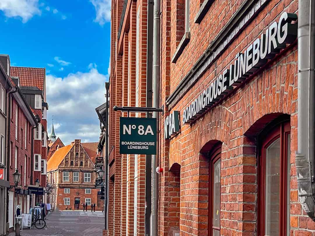 Eine schmale Straße in Lüneburg, Deutschland, gesäumt von roten Backsteinhäusern, darunter eine Pension mit dem Schild "N°8A BOARDINGHOUSE LÜNEBURG". Ein paar Menschen gehen in der Ferne unter einem teilweise bewölkten Himmel.