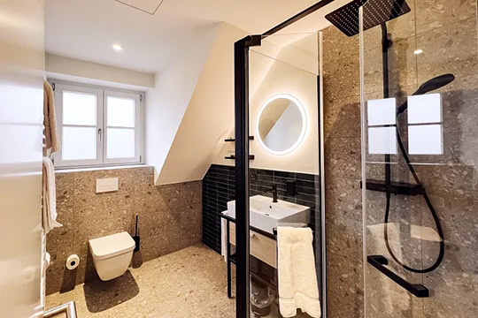 Modernes Badezimmer mit einer großen begehbaren Dusche, einer schwarz gerahmten Glastür, einer Wandtoilette, einem weißen Waschbecken mit rundem, hinterleuchtetem Spiegel und einem Fenster, das Tageslicht hereinlässt.