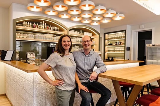 Eine Frau und ein Mann lächeln, während sie an einem hölzernen Bartresen in einer modernen, gut beleuchteten Bar oder einem Café mit Regalen voller Gläser und Flaschen im Hintergrund stehen und sitzen.