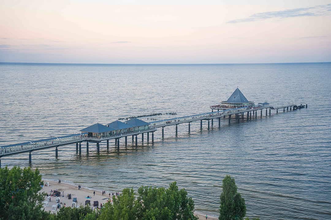 Ein langer Steg mit Pavillonstrukturen erstreckt sich von einem Sandstrand aus über das ruhige Meer, an dem Menschen entlang spazieren. Bäume und Besucher sind am Strand unter einem pastellfarbenen Himmel bei Sonnenuntergang zu sehen.