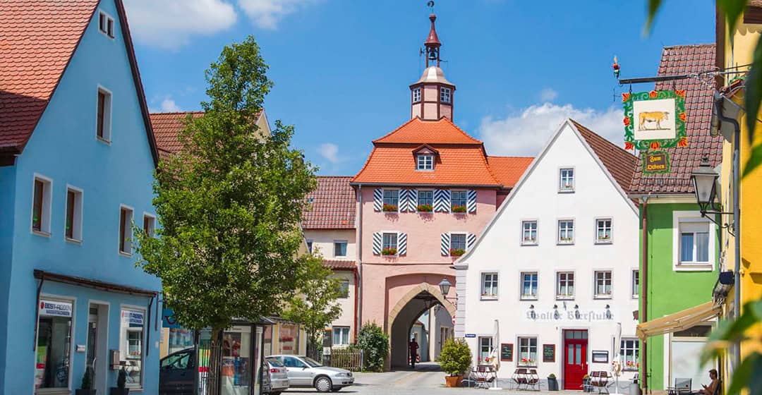 Ein malerisches europäisches Dorf mit farbenfrohen Gebäuden, einem zentralen Uhrenturm mit rotem Dach und bogenförmigem Tor, Sitzgelegenheiten im Freien und einem klaren, blauen Himmel über sich.