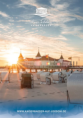 Ein malerischer Strand bei Sonnenuntergang mit Strandkörben im Sand, einer Seebrücke mit historischen Gebäuden und wehenden Fahnen im Wind auf der Insel Usedom, Deutschland. Der Himmel ist teilweise bewölkt, aber die Sonne scheint durch.