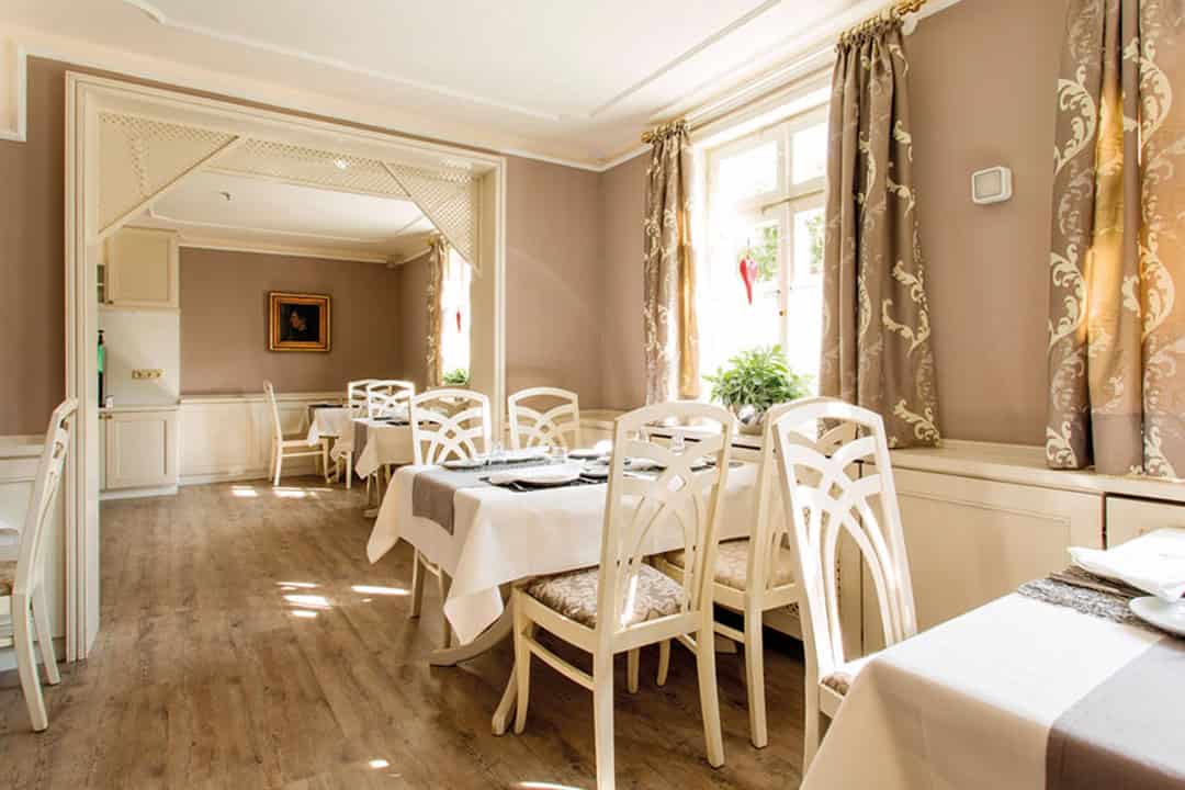 Ein heller, eleganter Speisesaal mit hellen Holzböden, cremefarbenen Wänden, weißen Stühlen und mit weißen Tischdecken bedeckten Tischen. Das Sonnenlicht strömt durch große Fenster mit drapierten Vorhängen herein.