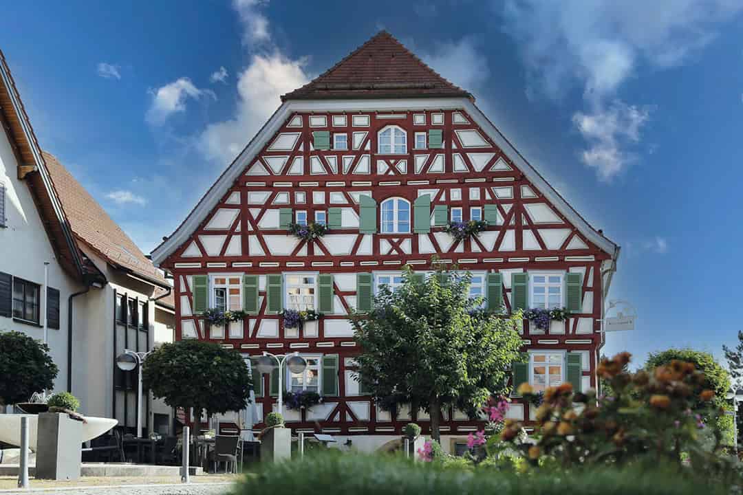 Ein traditionelles deutsches Fachwerkhaus mit rot-weißer Fassade, grünen Fensterläden und Blumenkästen an den Fenstern, vor einem blauen Himmel mit vereinzelten Wolken und umgeben von Bäumen und Blumen.