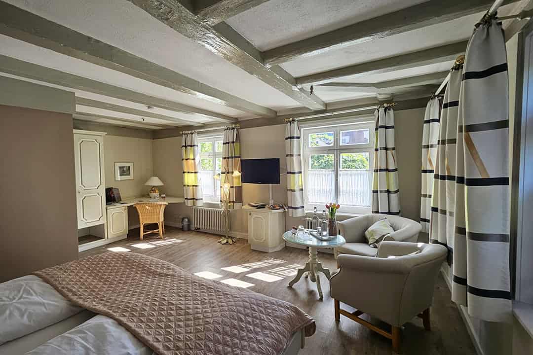 Gemütliches, sonnendurchflutetes Hotelzimmer mit Holzbalken, Doppelbett, kleinem Tisch, Sesseln, Fernseher, Korbtisch und großen Fenstern mit gestreiften Vorhängen. Helle, neutrale Töne und eine weiche, einladende Einrichtung.