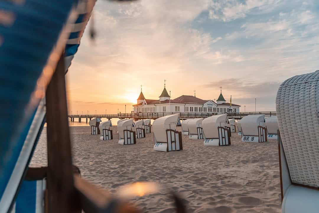 Sonnenaufgang über einem Sandstrand mit weißen Strandkörben, die dem Meer zugewandt sind. Im Hintergrund ragt ein Pier mit einem historischen Gebäude mit begrünten Türmen unter einem sanften, farbigen Himmel ins Wasser.