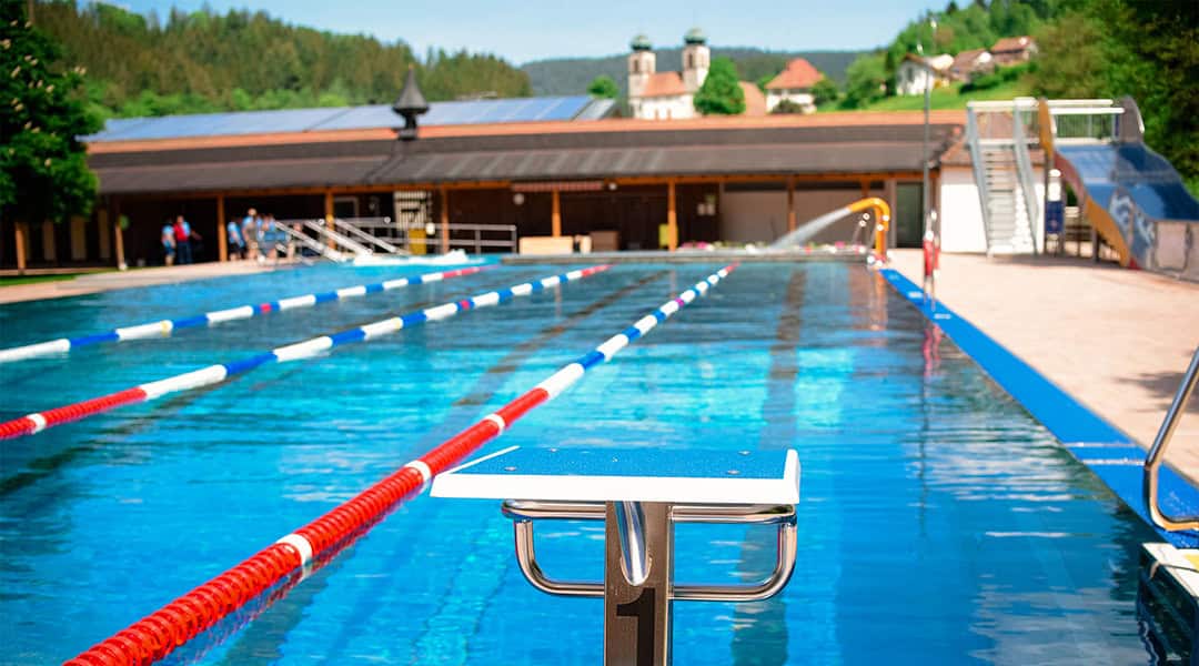 Ein Schwimmbad mit Bahntrennern und einem Startblock im Vordergrund, umgeben von Bäumen und einem Gebäude mit einem Schrägdach. Im Hintergrund sind Hügel und eine Kirche zu sehen.