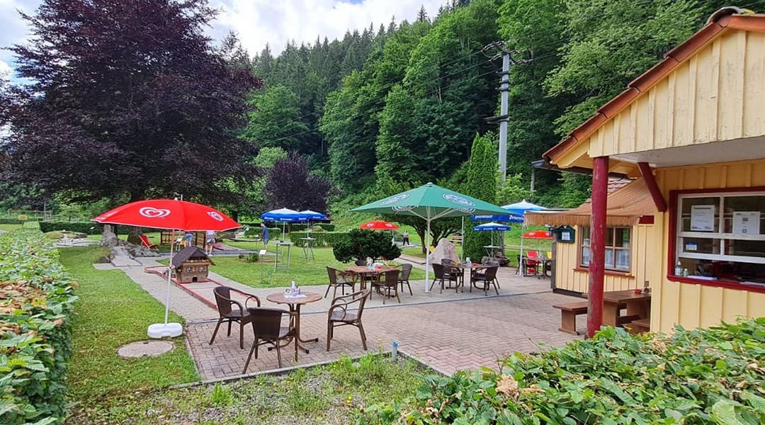 Außencafé mit Tischen und Stühlen unter bunten Sonnenschirmen auf einer Terrasse, umgeben von Grün und Bäumen. Auf der rechten Seite steht ein gelbes Gebäude, und im Hintergrund erhebt sich ein bewaldeter Hügel.