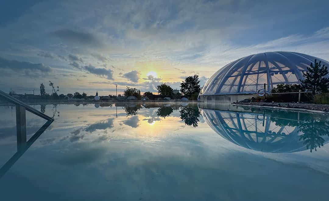 Zwei große Raubvögel mit dunklem Gefieder fliegen über einer Steinmauer vor einem hellblauen Himmel, die Flügel weit ausgebreitet und die Krallen sichtbar.