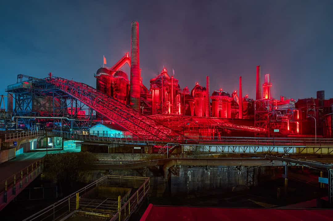 Zwei große Raubvögel mit dunklem Gefieder fliegen über einer Steinmauer vor einem hellblauen Himmel, die Flügel weit ausgebreitet und die Krallen sichtbar.