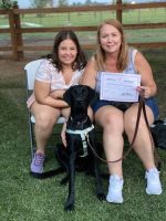 Certified therapy dog with young girl and woman, holding achievement certificate at High Plains K-9 Connections outdoor event, promoting service dog training and canine therapy programs.