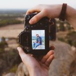 A person taking a photo of a man sitting outdoors on a rock, capturing a scenic landscape with a DSLR camera.