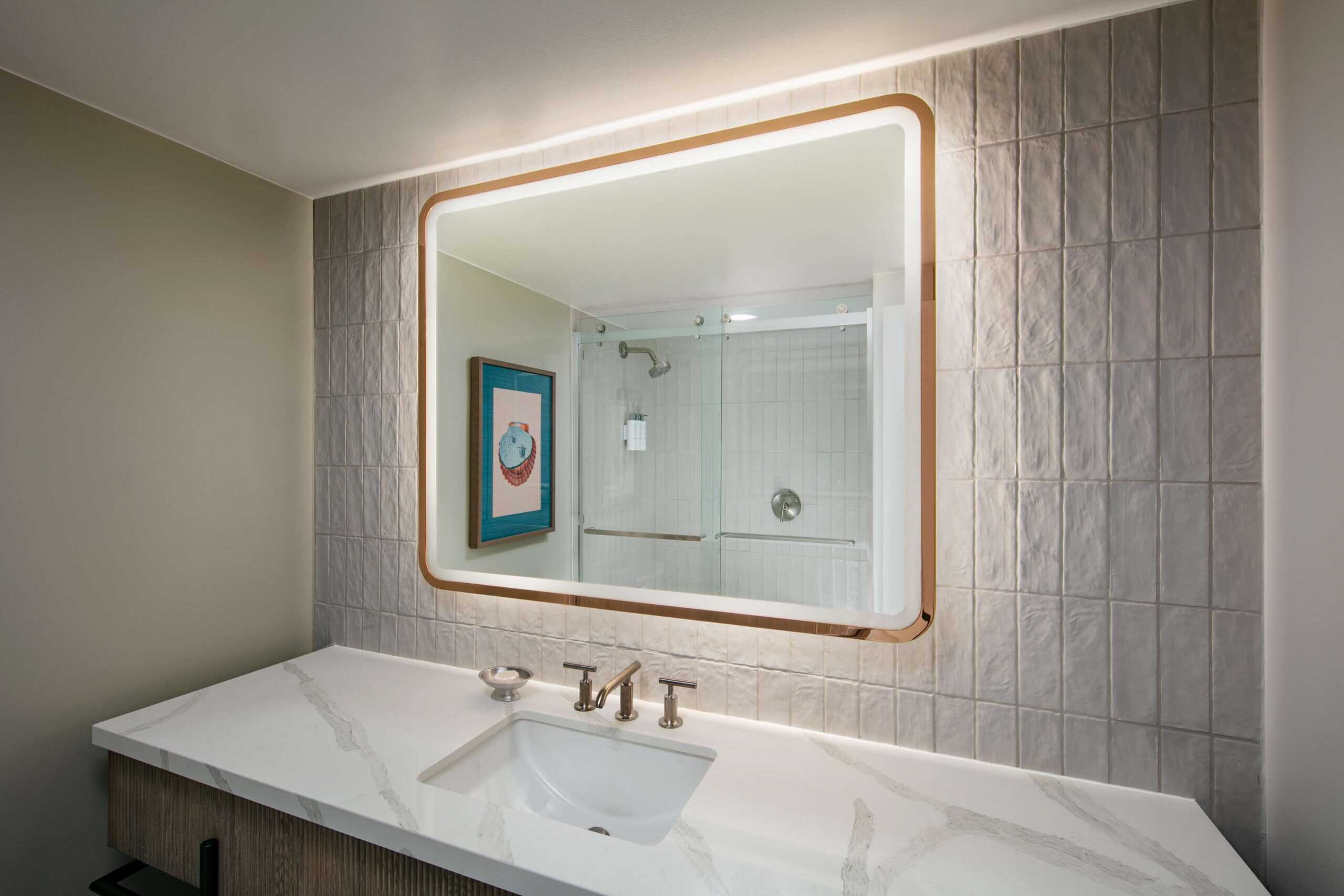 Bathroom vanity with a lit mirror and a shower in the background.