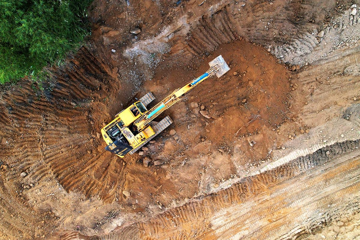 Aus der Vogelperspektive ist ein gelber Bagger zu sehen, der inmitten einer tiefen Baugrube Erde aushebt. Die Maschine arbeitet präzise im beengten Raum, umgeben von senkrechten Erdwänden. Das Bild zeigt eindrucksvoll die Dimension und Tiefe des Aushubs sowie den Einsatz moderner Baumaschinen bei Erdarbeiten auf einer Baustelle. Vogelperspektive auf gelben Bagger bei Erdarbeiten in tiefem Baugrubenloch.