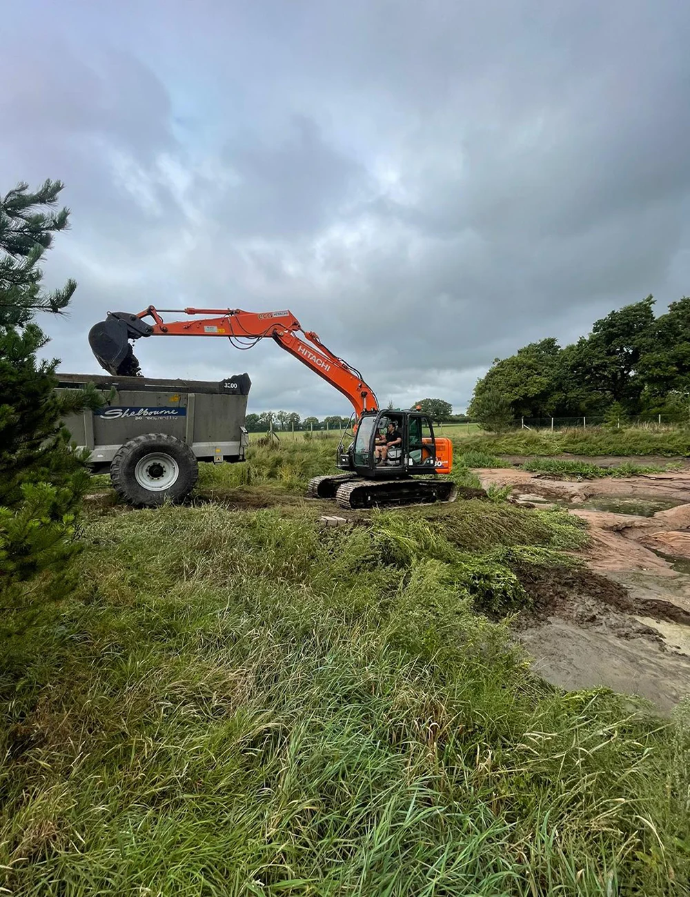 Digger and rear discharge spreader Excavator loading dump truck in grassy field