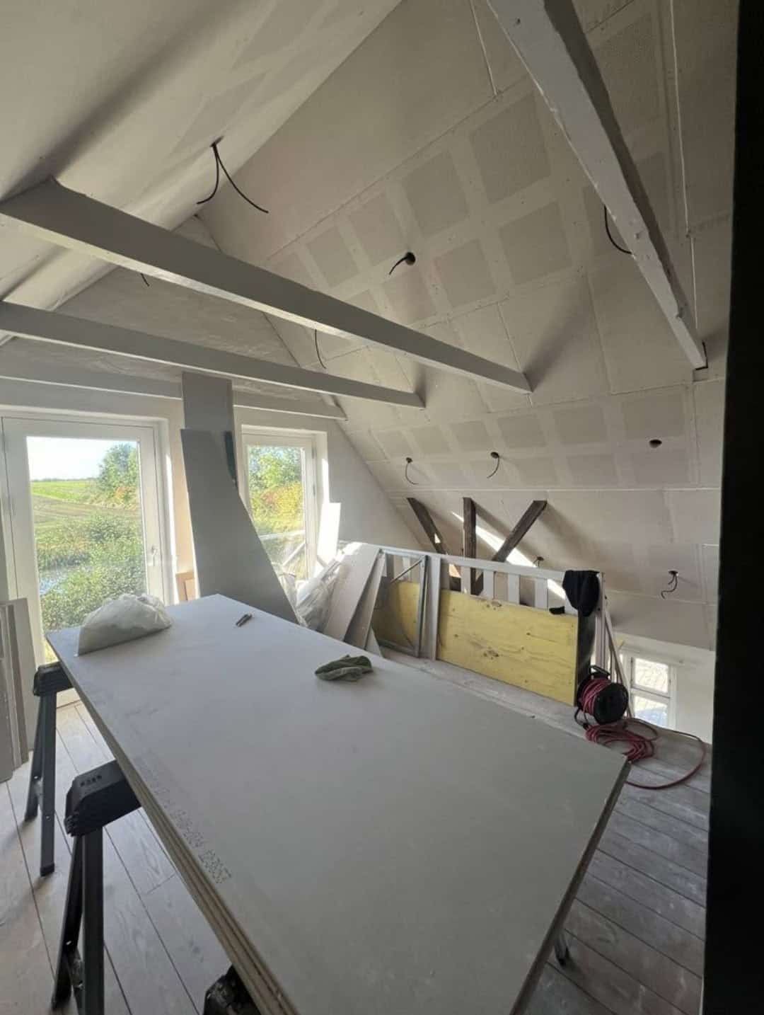 Ufærdigt rum med skråloft og et stort bord med tegningsmateriale.