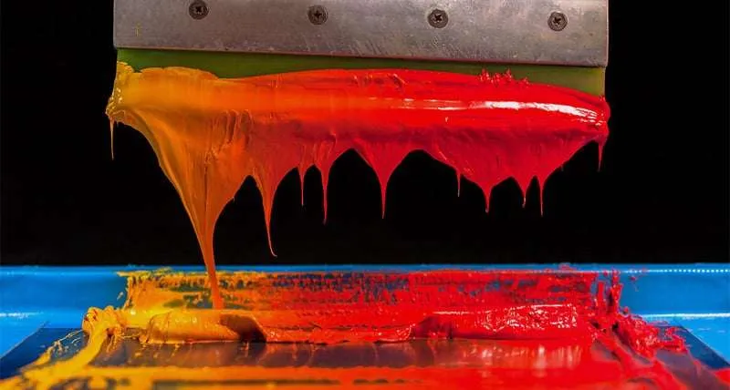 Close-up of a screen printing squeegee applying red and orange ink on a screen in Jacinto City, highlighting vibrant colors and printing technique.