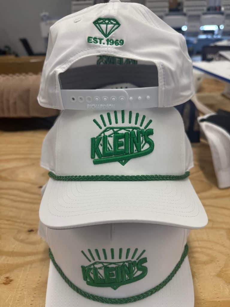 White Klein's cap with green logo and sunburst design, adjustable strap, and embroidered details.