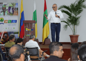 Hombre presentando en evento sobre conservación de recursos naturales en Huila, con banderas de Colombia y Huila, y audiencia atenta.