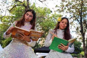 Conjunto de dos jóvenes leyendo libros en un entorno natural en Huila Tv, celebración cultural y artística en Colombia, con vestidos brillantes y naturaleza alrededor.