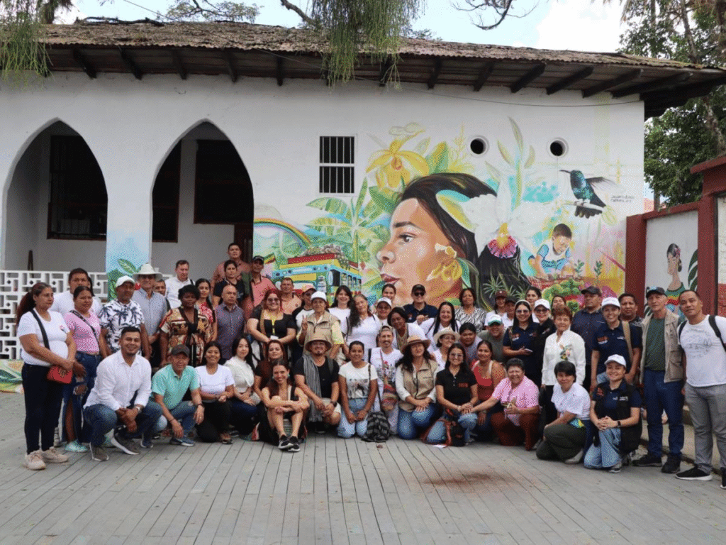 Diversos miembros de la comunidad posan frente a un mural artístico en Huila, Colombia, destacando la cultura, naturaleza y desarrollo social del departamento en una imagen que representa identidad y unión.